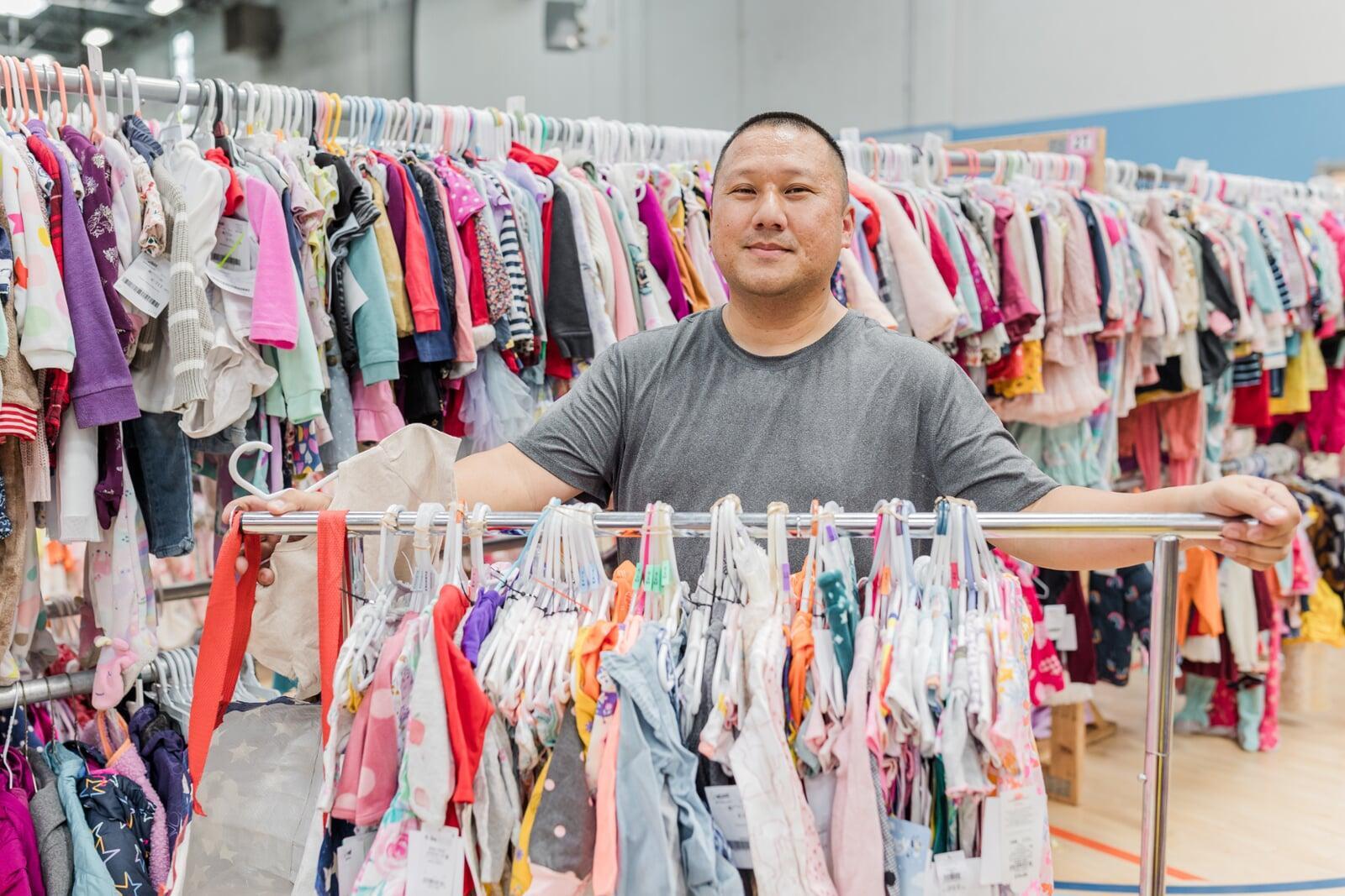 A woman kneels in front of racks of boys clothing. She is hold hangers full of boys clothing in both hands.